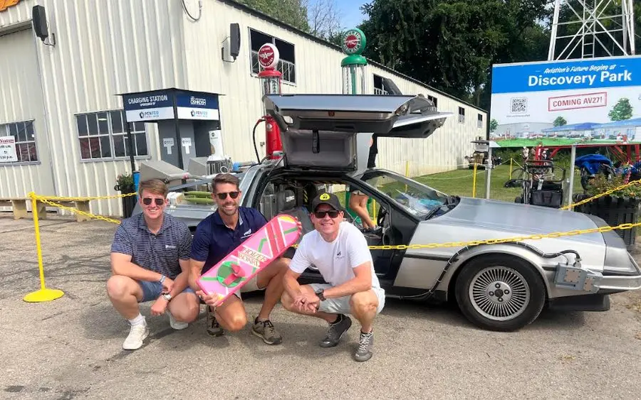 Three members of the Peregrine Howard team crouching by a DeLorean at EAA AirVenture Oshkosh